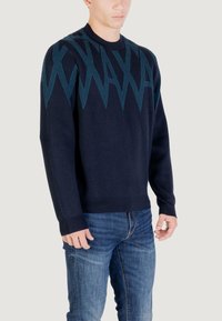 Homme portant un pull bleu marine avec des motifs en zigzag teal sur la poitrine et les épaules, associé à un jean bleu.