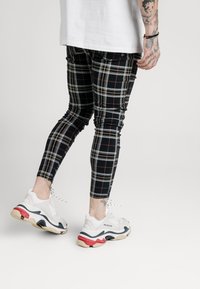 Person klädd i svart, vitt och rött rutigt byxor samt vita klumpiga sneakers med svarta och röda detaljer, står framför en enkel ljus bakgrund.