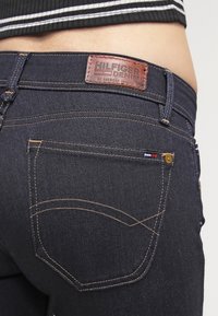Vaqueros de denim oscuro con una etiqueta de cuero "HILFIGER DENIM" en la cinturilla, costuras amarillas contrastantes y un pequeño acento tricolor en el bolsillo.