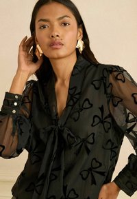 Černá průsvitná halenka se vzory mašlí, zavazováním na krku a nadýchanými rukávy. Knoflíkové detaily na manžetě s mírným leskem látky.