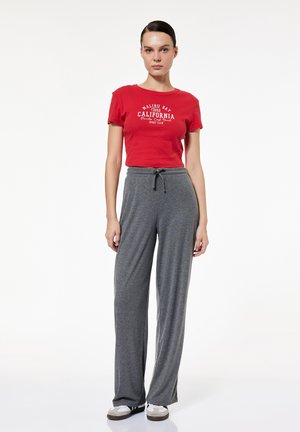 Mujer de pie con una camiseta corta roja con estampado de California, pantalones grises con cordón y zapatillas blancas sobre fondo blanco.