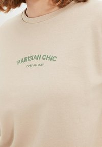 Sudadera beige hecha de tela suave con escote redondo y texto verde que dice "PARISIAN CHIC POSE ALL DAY." Textura suave.