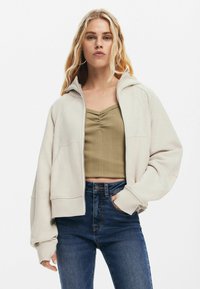 Ljusbeige zip-up huvtröja med huva, ribbad struktur, två framfickor och vida ärmar, i kombination med en mörkblå högmidjad kjol.