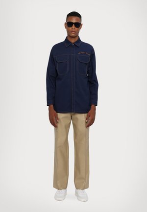Navy jacket with two front pockets and visible stitching, paired with beige wide-leg pants and white sneakers. Model wears dark sunglasses.