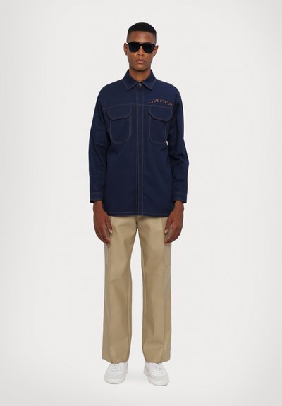 Giacca navy con due tasche frontali e cuciture in vista, abbinata a pantaloni beige a gamba larga e sneakers bianche. Il modello indossa occhiali da sole scuri.
