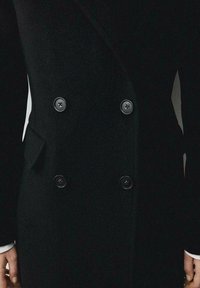 Palton dublu încheiat, de culoare neagră, realizat dintr-un material texturat, având butoane mari negre și un design elegant cu o croială slim și rever tăiat.