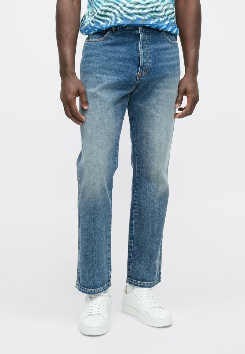 Jean droit bleu clair en denim, avec un effet délavé sur les cuisses et un design classique à cinq poches. Porté avec des baskets blanches.