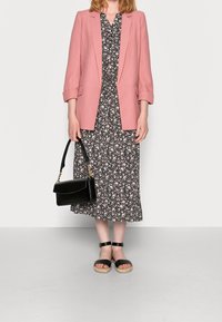 Roze blazer over een zwarte bloemenjurk, met witte en roze bloemen. Zwarte schoudertas met gouden hardware en zwarte platformsandalen.