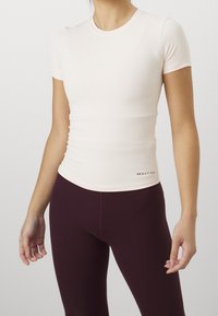 Vaaleanpunainen kiinteä t-paita, jossa on lyhyet hihat, sileä pinta, pyöreä pääntie ja pieni logo vasemmassa alakulmassa. Yhdistetty tummiin burgundinvärisiin legginsseihin.