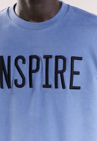 Jasnoniebieska bawełniana koszulka z dużymi czarnymi haftowanymi literami, które na piersi tworzą napis "INSPIRUJ". Prosty krój z dekoltem w serek.