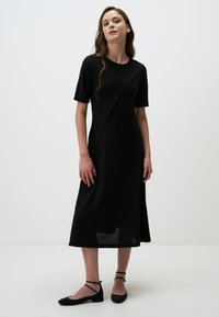 Robe noire mi-longue avec des manches courtes, tissu texturé, design asymétrique et fentes latérales; assortie avec des chaussures plates noires à brides de cheville.