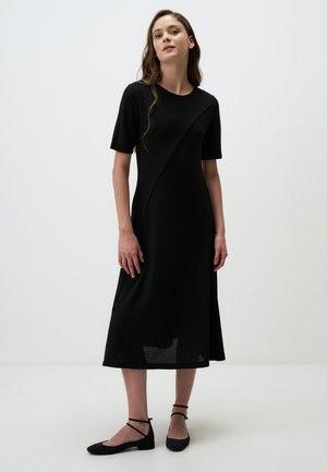 Black midi dress with short sleeves, textured fabric, asymmetrical design, and side slits; paired with black flat shoes with ankle straps.