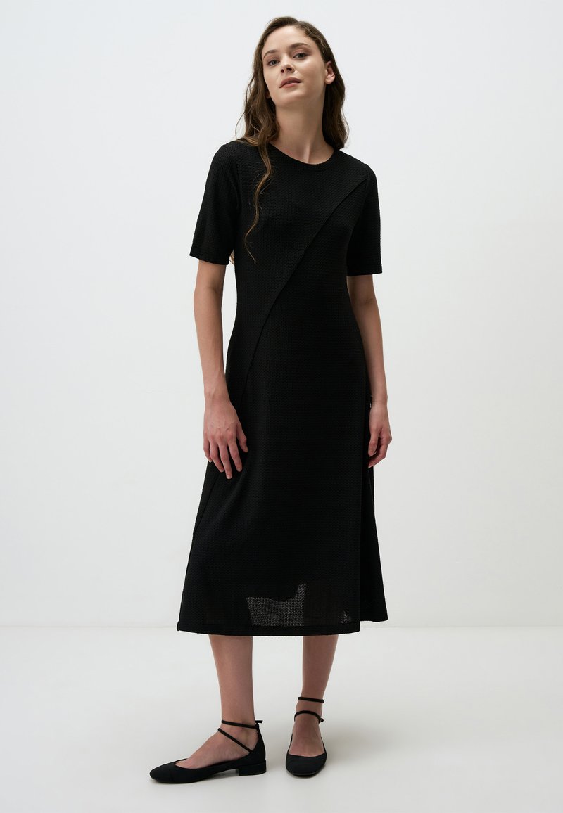 Robe noire mi-longue avec des manches courtes, tissu texturé, design asymétrique et fentes latérales; assortie avec des chaussures plates noires à brides de cheville.