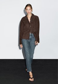 Bruine suède overhemdjack met een kraag en knoopsluiting, gecombineerd met lichtblauwe rechte jeans en bruine sandalen. Effen witte achtergrond.