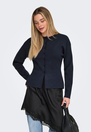 Piklikeste blondide juustega naine kannab tumesinist nööbitavat kardigani, musta asümmeetrilist pitsäärisega seelikut hele siniste teksade peal, hoides musta käekotti.