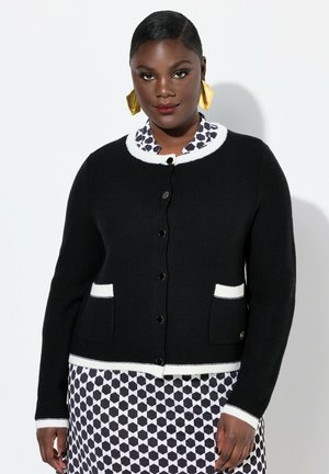 Cardigan noir avec des bordures blanches, en tissu texturé, col rond, fermeture à boutons et deux poches avant. Porté sur une robe à motifs.