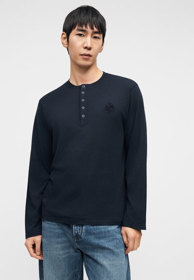 Μπλε ναυτικού χρώματος μπλουζάκι με μακριά μανίκια, στρογγυλή λαιμόκοψη και κούμπωμα με πέντε κουμπιά. Διαθέτει κεντημένο λογότυπο στο στήθος. Μαλακό βαμβακερό ύφασμα.