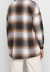 Osoba oblečená v hnědo-bílo-černé kostkované bundě s dlouhými rukávy a světlými kalhotami, stojící před jednobarevným pozadím.