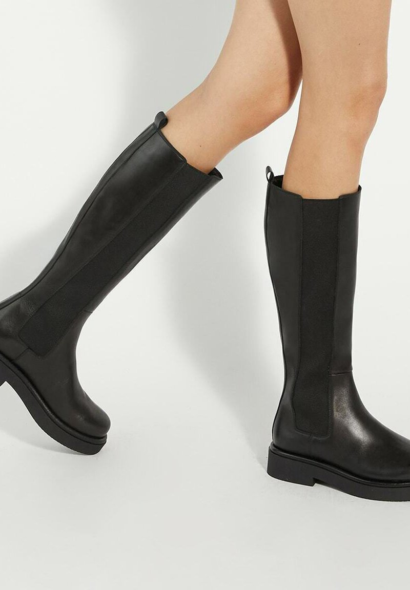 Zwarte kniehoge laarzen met een gladde leren afwerking, elastische zijpanelen en een dikke rubberen zool. Voorzien van trekriemen aan de bovenkant voor gemakkelijk aan- en uittrekken.