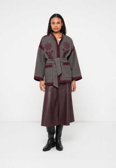 Γκρίζα ζακέτα από μάλλινο υλικό με μαρτούν εμπριμέ λουλούδια, ζωνάκι στη μέση και φαρδιά μανίκια, συνδυασμένη με φούστα από μπορντό δέρμα και μαύρες μπότες μέχρι το γόνατο.