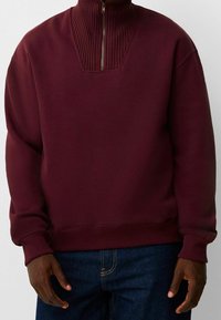 Sudadera burdeos con cremallera en el cuello, textura acanalada en el cuello, corte relajado y puños acanalados, combinada con jeans de mezclilla oscura.