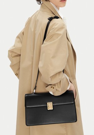 Person trägt einen beigen Trenchcoat und hält eine schwarze Leder-Umhängetasche mit goldfarbenem Verschluss mit einer Hand in der Manteltasche.