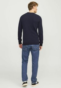 Navy knitted sweater with round neckline, paired with blue denim jeans and black sneakers with white sole and blue accents, viewed from the back.