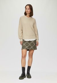 Beige gestreifter Pullover mit überschnittenen Schultern, über einem weißen Kragenshirt getragen. Grüner Karorock mit einem Saum knapp über dem Knie, kombiniert mit schwarzen Knöchelstiefeln.