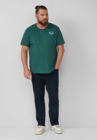 Groene gemêleerde katoenen t-shirt met korte mouwen, ronde halslijn en kleine witte grafische tekst op de borst, gecombineerd met donkere jeans en sneakers.