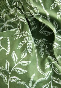 Tessuto verde con motivo a foglie bianche, caratterizzato da diverse forme di foglie e dettagli raffinati. Include una tasca laterale e una texture morbida.