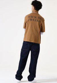 Camisa de algodón marrón de manga corta con "VOLVER EN 2 SEMANAS" impreso en la parte de atrás, combinada con pantalones de mezclilla azul oscuro y zapatillas blancas.