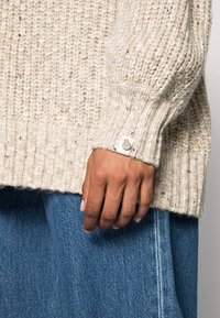 Virkad tröja i ljus beige med texturerat mönster och ribbade muddar. Inkluderar en liten vit etikett med logotypdetalj. Bärs med blå denim.