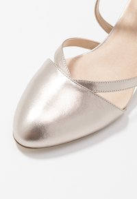 Metalická stříbrná botka na vysokém podpatku se špičatou špičkou, vybavená kříženými pásky a hladkou texturou. Minimalistický design, neutrální barva.