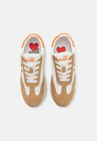 Tan and white sneakers with suede and mesh, featuring orange accents, round laces, and a red heart logo with “LOVE MOSCHINO” on the tongue.