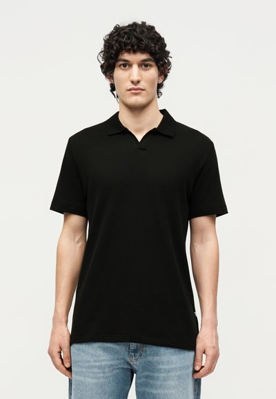 Jeune homme aux cheveux bouclés, portant un polo noir à manches courtes et un jean bleu clair, debout sur un fond uni.
