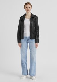 Schwarze Lederjacke mit Reißverschluss und schrägem Kragen, getragen über einem weißen Tanktop. Hellblaue, ausgestellte Jeans und weiße Sneaker runden das Outfit ab.