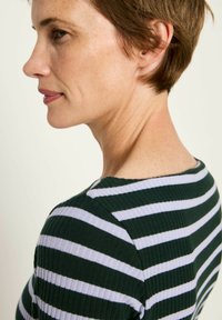 Frau mit kurzen braunen Haaren, die ein gestreiftes Oberteil in Dunkelgrün und Weiß trägt, von links vor einem einfachen hellen Hintergrund.