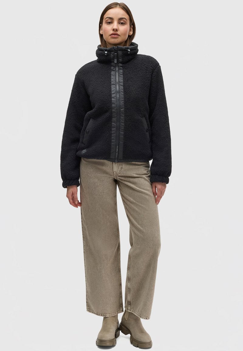 Schwarze Fleecejacke mit hohem Kragen, strukturierter Oberfläche und Frontreißverschluss. Kombiniert mit hellbraunen weiten Hosen und beigen Knöchelstiefeln.
