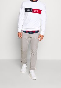 Sudadera blanca con logo "HILFIGER" en azul y rojo, sobre una camisa de cuadros. Chinos beige y zapatillas blancas completan el conjunto.