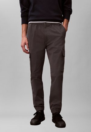 Mężczyzna ubrany w ciemnoszare joggery cargo z elastycznymi ściągaczami i czarne trampki, stoi z jedną ręką w kieszeni.