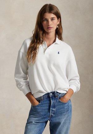 Kobieta z długimi brązowymi włosami, ubrana w białą, długorękawową koszulkę polo i niebieskie dżinsy, z rękami w kieszeniach na jednolitym tle.