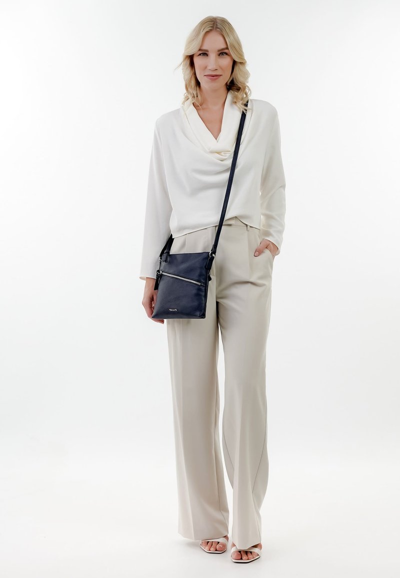 Blondīne sieviete baltā blūzē, bēšās platām biksēm, baltās papēžu sandalēs un melnu krustenes tipa somiņu, stāvot pret baltu fonu.