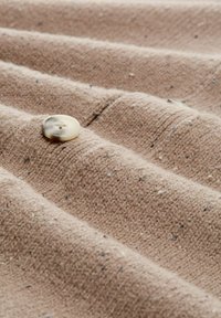 Beiger Stoff mit strukturiertem Gewebe, in Falten drapiert und mit kleinen schwarzen Punkten verziert. Ein hellfarbener Knopf sitzt auffällig auf dem Stoff.