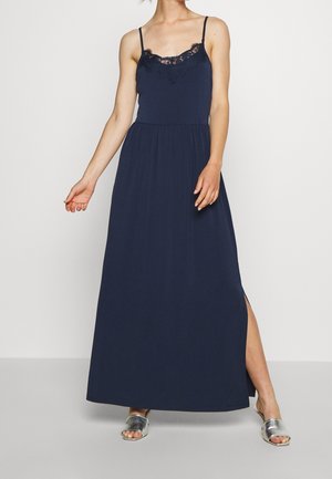 Marineblaues Maxikleid mit Spitzenbesatz am Ausschnitt, figurbetontem Oberteil, ausgestelltem Rock und seitlichem Schlitz; kombiniert mit silbernen Slide-Sandalen.