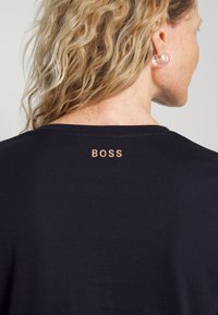 Svart t-shirt med rund halsringning och korta ärmar. "BOSS"-logotypen visas i ljusbrunt på baksidan nära halsringningen. Mjuk tygstruktur.