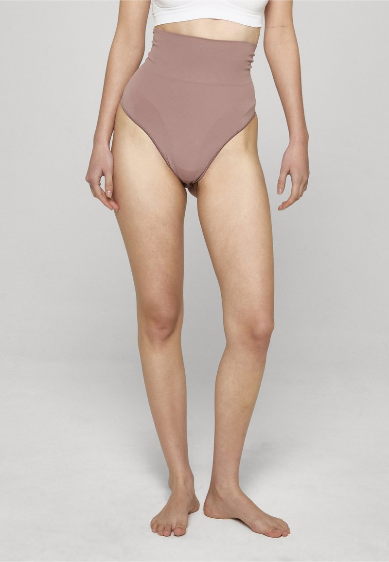 Moteris dėvi aukšto liemens alyvinės spalvos korpusinius apatinukus ir baltą trumpą marškinėlį, basomis stovinti priešais paprastą šviesiai pilką foną.