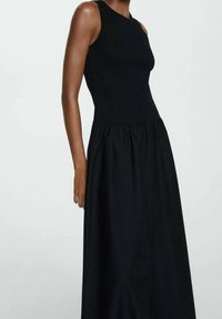 Robe noire sans manches avec un corsage ajusté et une jupe froncée. Confectionnée dans un tissu lisse, elle présente un décolleté rond et une coupe longue.