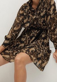 Musta sifonimekko, jossa on kultaisista sävyistä koostuva paisley-kuvio, pitkät hihat ja rento, kiristetty vyötärösuunnittelu.