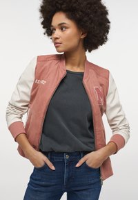 Jasnoróżowa kurtka bombowiec z kremowymi rękawami, ściągaczami i zamkiem. Posiada haftowane oznaczenie "EST 32" oraz teksturowaną literę "M". Nosi się ją na ciemnej koszuli.