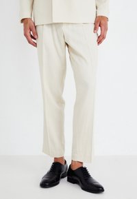 Pantalones de vestir beige confeccionados en un tejido suave, con corte recto y pliegues marcados, acompañados de zapatos de cordones negros pulidos.